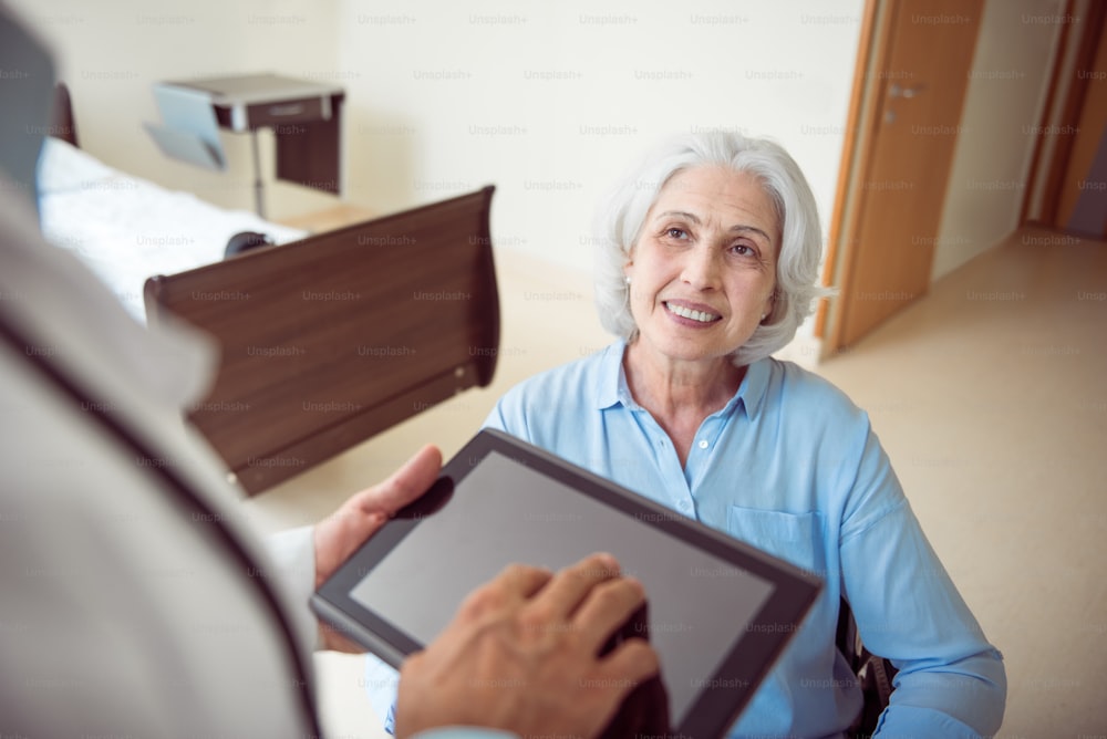 Beste Klinik. Abgeschnittenes Bild einer glücklichen und lächelnden Frau im Rollstuhl und eines Arztes mit digitalem Tablet