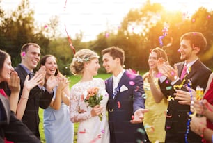 Full length portrait of newlywed couple and their friends at the wedding party showered with confetti in green sunny park
