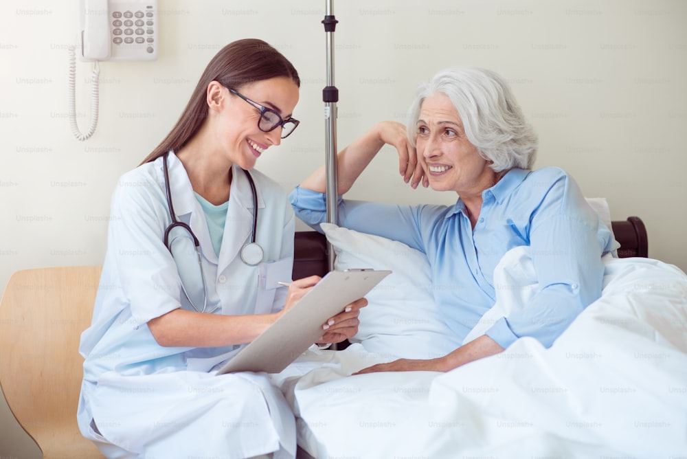 Important job. Delighted senior woman lying in bed and doctor sitting near her and writing something down with clipboard in hands