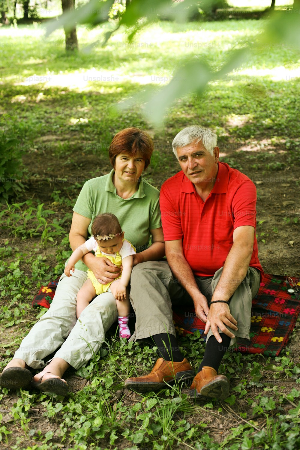 Grandparents sitting on grass with baby girl in the park. Above view. Looking at camera.