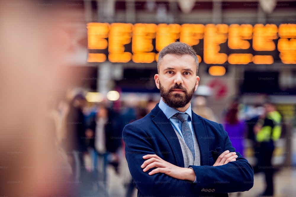 Uomo d'affari hipster in giacca e cravatta in attesa all'affollata stazione ferroviaria di Londra, braccia incrociate