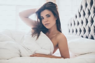 Beautiful young woman looking at camera and keeping hand in hair while lying in bed