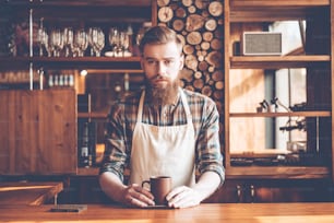 Young bearded man in apron looking at camera and holding coffee cup while standing at bar counter