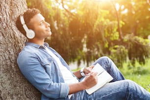 Attractive young man is relaxing in park. He is writing in notebook with inspiration and dreaming. The guy is sitting on grass and listening to music