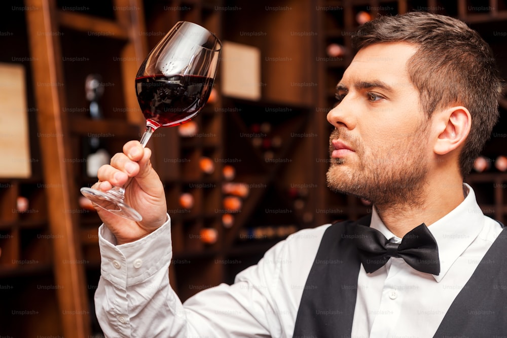 Confident male sommelier examining glass with wine while standing near the wooden shelf