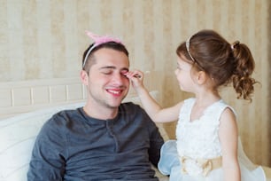 Cute little girl in princess dress putting on colorful make up on her father