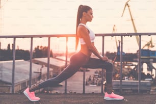 Full length side view of beautiful young woman in sports clothing doing stretching exercises while standing on the bridge with evening sunlight and urban view in the background