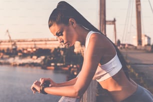 Hermosa joven con ropa deportiva mirando su reloj de pulsera mientras está de pie en el puente