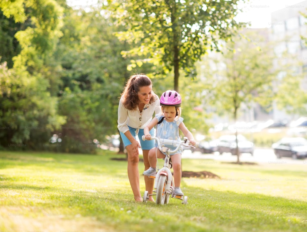 In a sunny city park, a mother gently pushes her blonde, blue-eyed daughter forward with encouragement as she learns to ride her bike. Thanks to her pink helmet, the daughter is practicing bike safety.