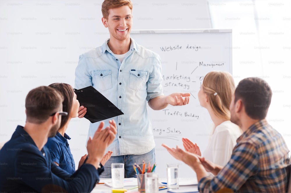 Group of business people in smart casual wear sitting together at the table and applauding to their colleague standing near whiteboard and smiling