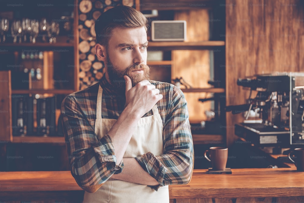Young bearded man in apron looking away and keeping hand on chin while standing at bar counter