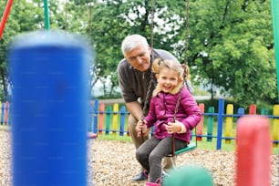 Caucasian grandfather pushing granddaughter on playground swing.