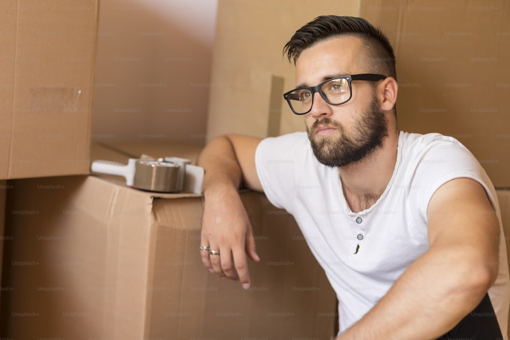 Young man moving in a new apartment, sitting on the floor, surrounded with cardboard boxes with packing machine placed on one of the boxes in the background