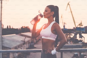 Beautiful young woman in sports clothing drinking water and looking tired while standing on the bridge with evening sunlight and urban view in the background