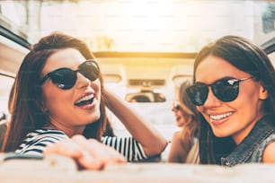Vista trasera de dos hermosas mujeres jóvenes y alegres que miran a la cámara con una sonrisa mientras están sentadas en el asiento trasero del automóvil