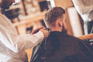 理髪店の椅子に座っている間、電気かみそりで美容師によって散髪される若いひげを生やした男の背面図の接写