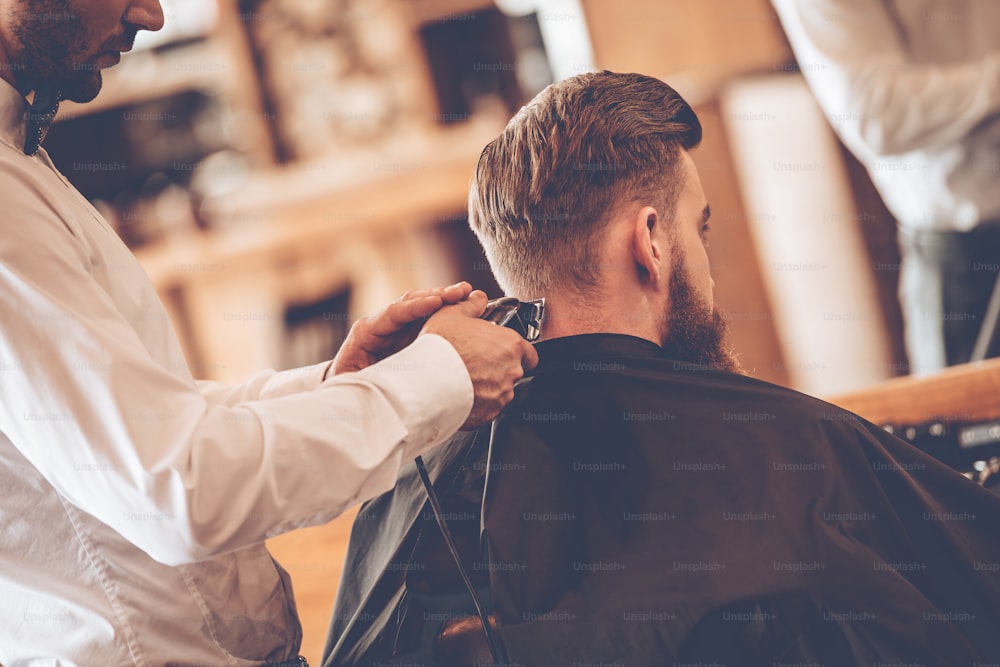 Close-up da vista traseira do jovem homem barbudo recebendo corte de cabelo pelo cabeleireiro com navalha elétrica enquanto sentado na cadeira na barbearia