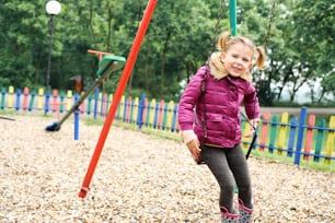 Cheerful little girl swinging on a playground in  park and looking at camera.