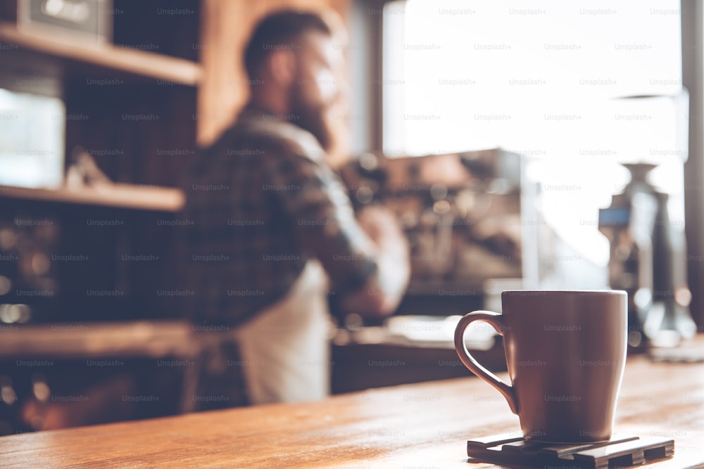 Focused picture of coffee cup standing at bar counter with young bearded man in apron making coffee in background