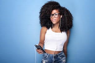 Hermosa mujer joven con afro escuchando música desde el teléfono móvil. Toma de estudio. Fondo azul.
