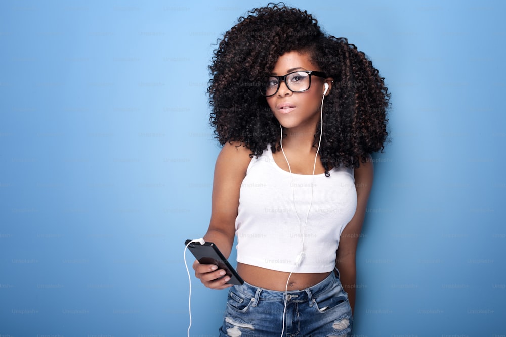 Schöne junge Frau mit Afro, die Musik vom Handy hört. Studioaufnahme. Blauer Hintergrund.