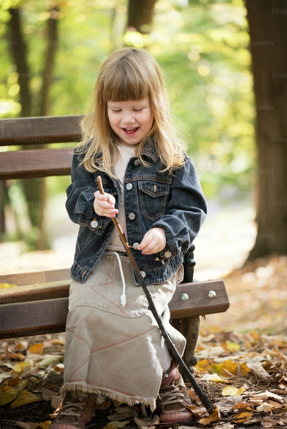 Beautiful little girl sitting alone on park bench and holding wooden stick. Looking down and smiling. Outdoor autumn portrait.