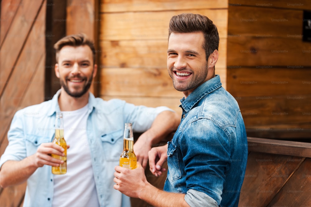 Zwei fröhliche junge Männer, die Flaschen mit Bier halten und in die Kamera schauen, während sie an der Bartheke lehnen
