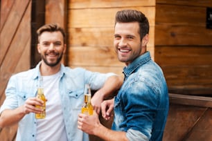 Deux jeunes hommes joyeux tenant des bouteilles de bière et regardant la caméra tout en se penchant au comptoir du bar