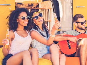 Two cheerful young women holding bottles with beer and smiling while man playing the guitar with minivan in the background