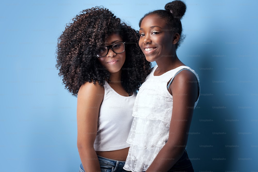 Dos hermosas chicas afroamericanas sonriendo, mirando a la cámara. Hermanas posando sobre fondo azul. Toma de estudio.