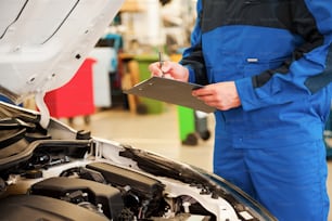 Close-up of man in uniform examining car and writing something in clipboard while standing in workshop