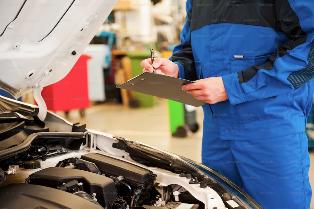 Close-up of man in uniform examining car and writing something in clipboard while standing in workshop