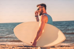 Joven guapo sosteniendo una tabla de skimboard y ajustándose las gafas mientras camina por la playa