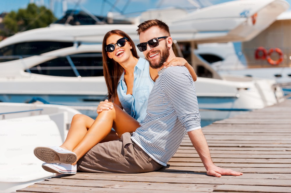 Cheerful young couple smiling and looking at camera while sitting on wooden pier