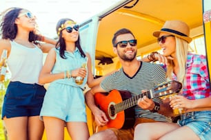 Joven guapo sentado en una minivan y tocando la guitarra mientras tres chicas están de pie cerca de él y sonríen
