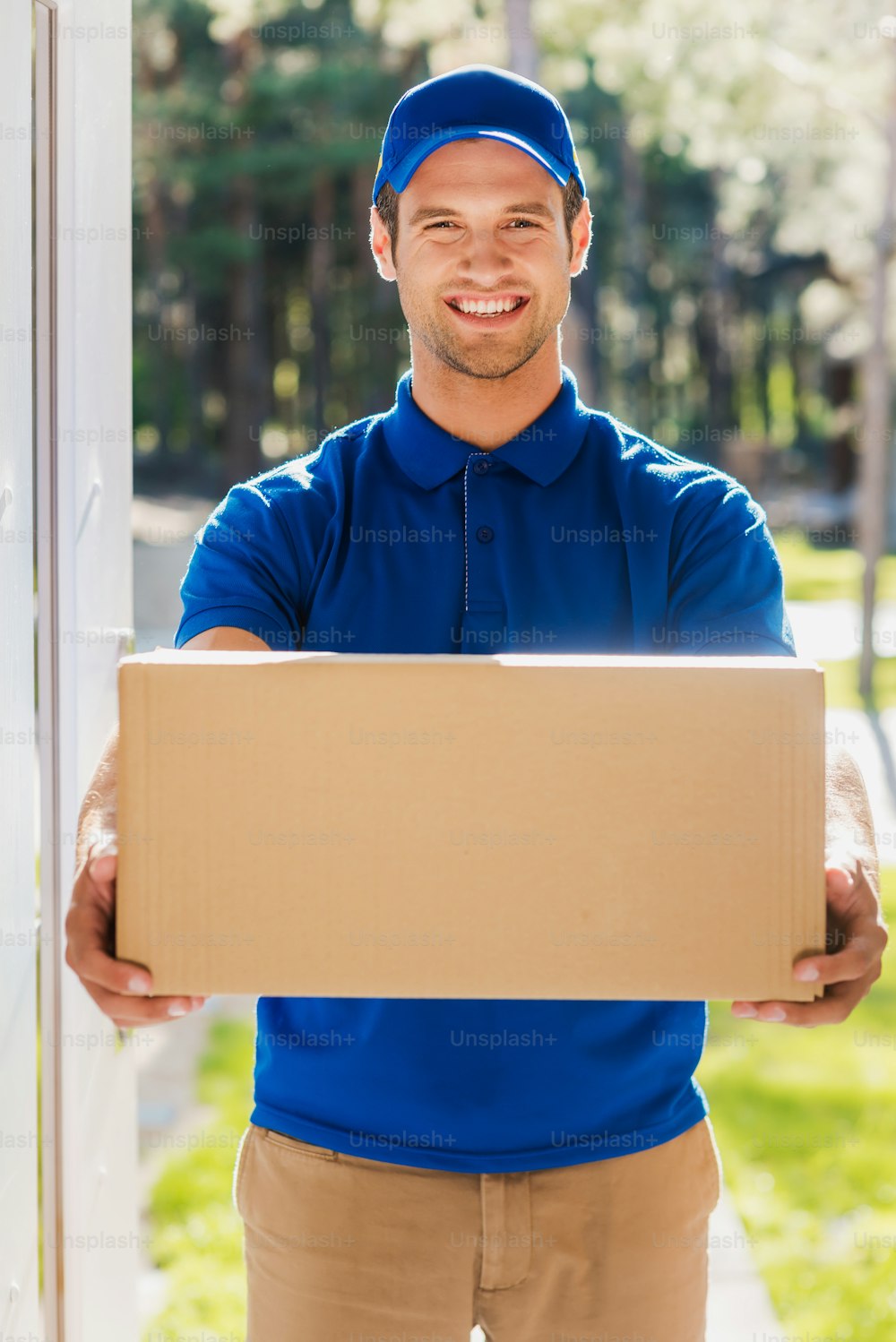 Joven repartidor feliz estirando una caja de cartón mientras está de pie en la entrada del apartamento