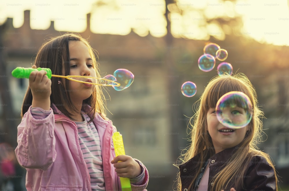 Emotionales Außenfoto von zwei kleinen Schwestern. Junge Mädchen haben eine gute Zeit im Park, blasen Blasen und lächeln.