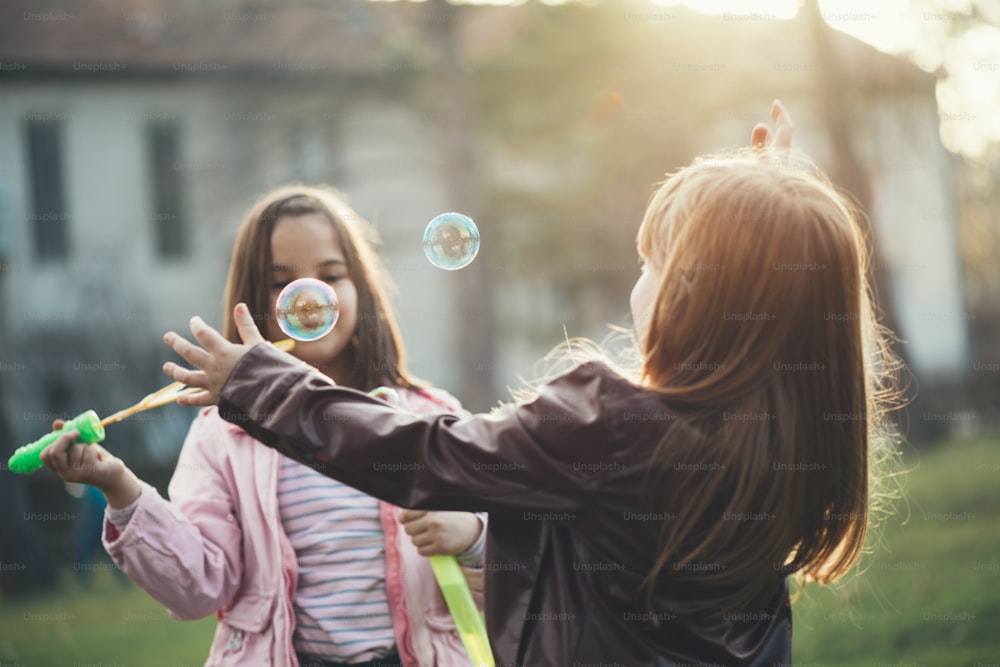 두 여동생의 감동적인 야외 사진. 어린 소녀들은 공원에서 즐거운 시간을 보내고 거품을 불고 웃고 있습니다.