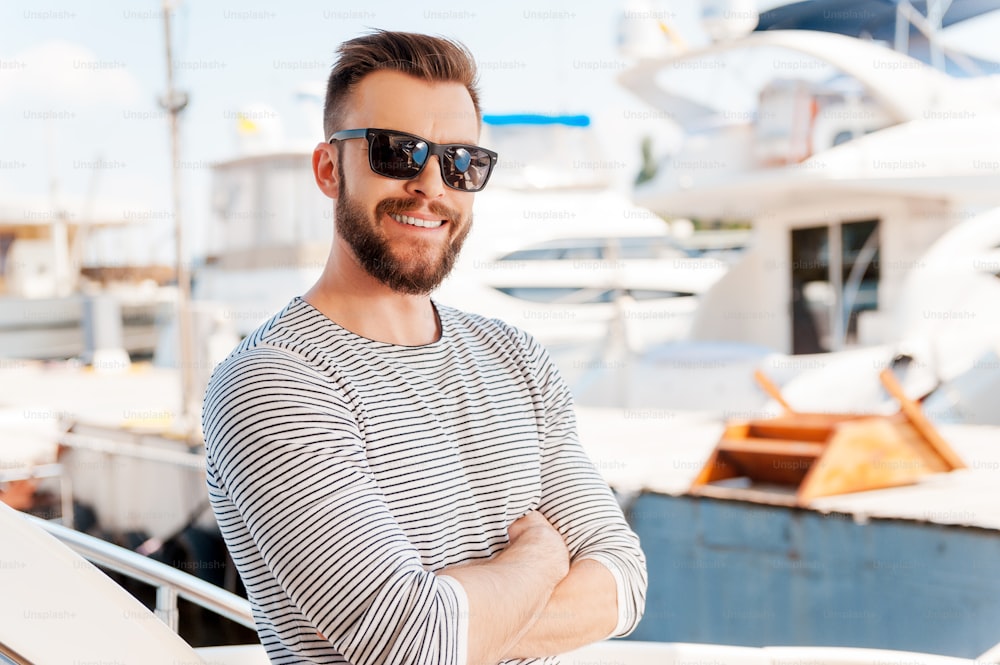 Smiling young man keeping arms crossed and looking at camera while standing on the board of yacht