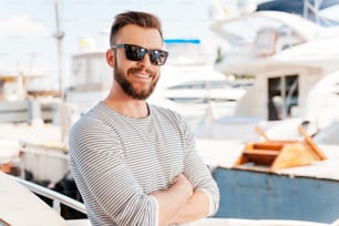 Lächelnder junger Mann, der die Arme verschränkt hält und in die Kamera schaut, während er an Bord einer Yacht steht