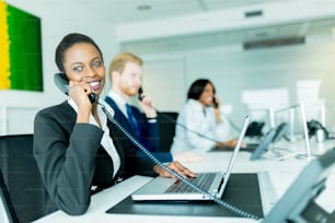 Una hermosa mujer joven y negra que trabaja en un centro de llamadas en una oficina con su pareja pelirroja al otro lado del escritorio hablando con otro cliente