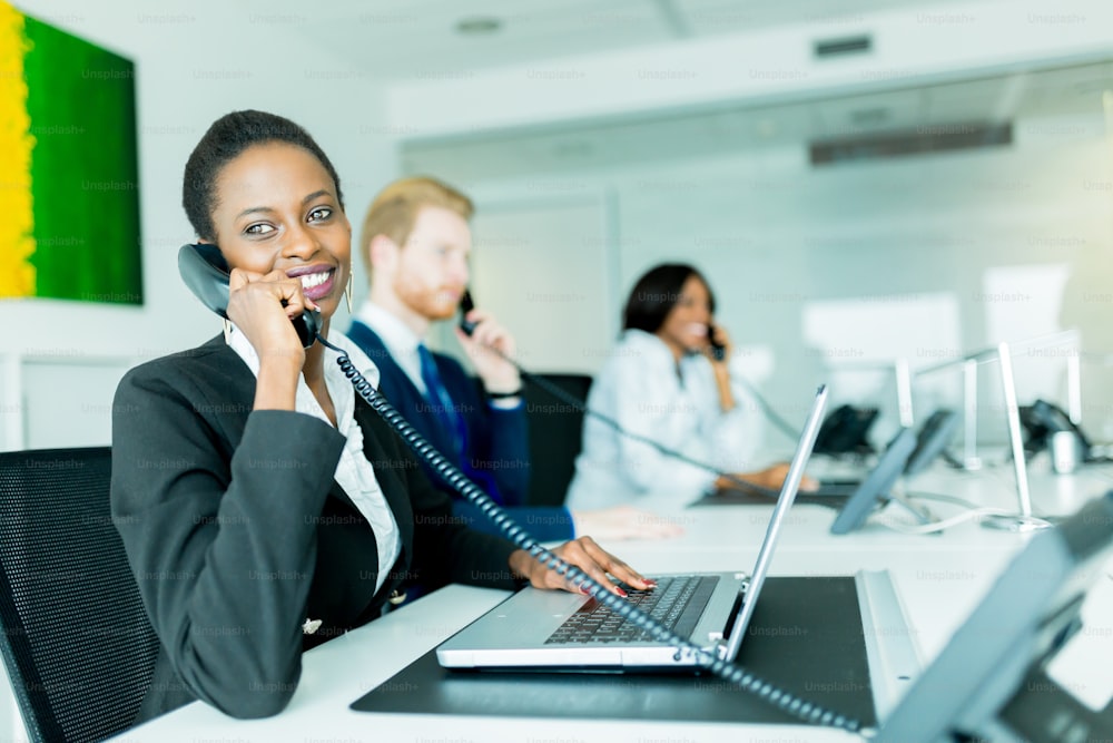 A beautiful, black, young woman working at a call center in an office with her red haird partner on the other end of the desk talking to another customer