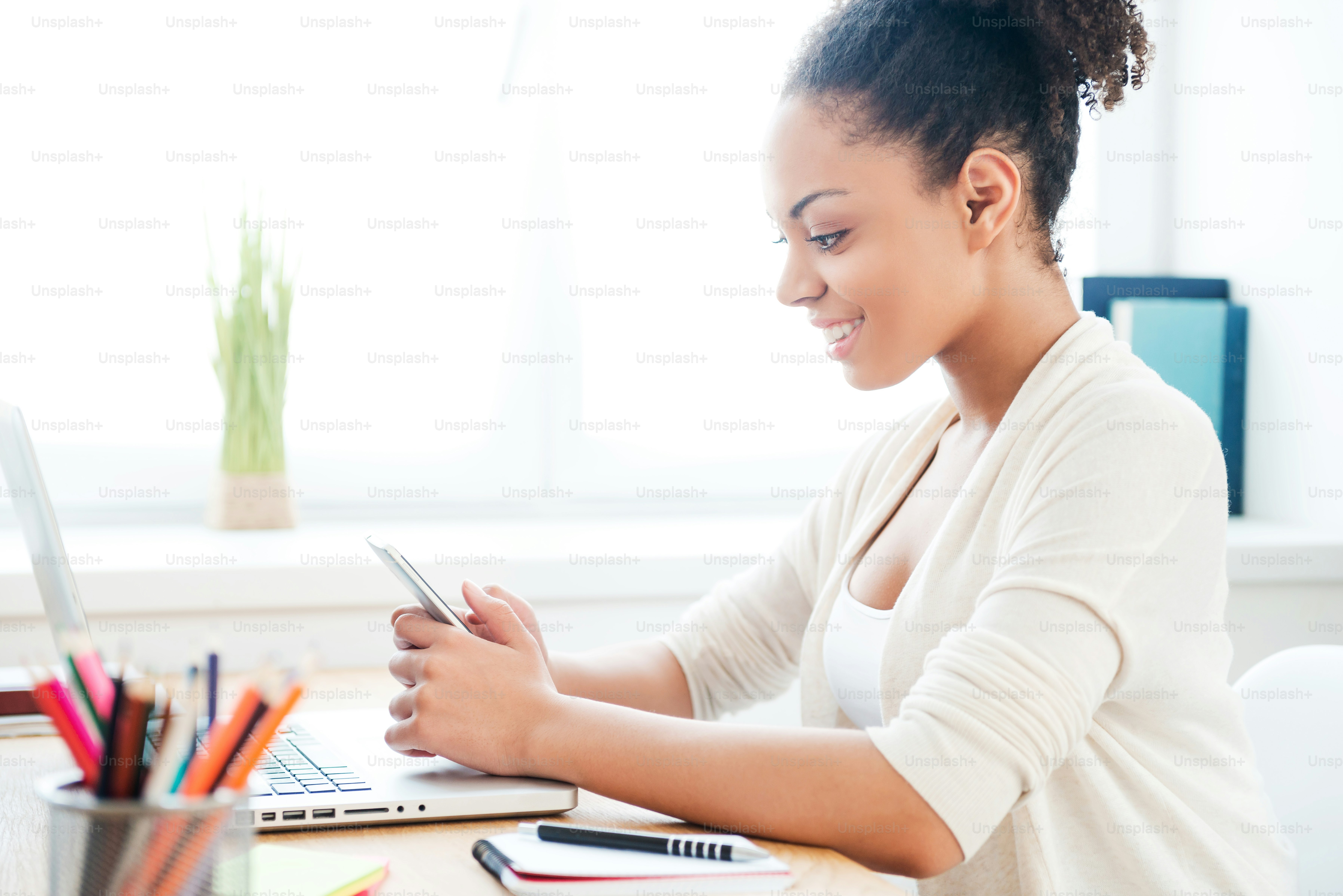 Tampilan samping wanita muda Afrika yang cantik memegang ponsel dan tersenyum sambil duduk di tempat kerjanya di kantor