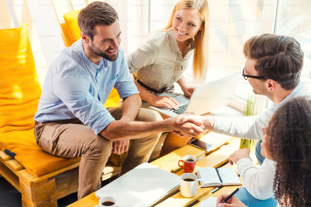 Two cheerful young men sitting at the wooden desk in office and shaking hands while two beautiful women looking at them and smiling