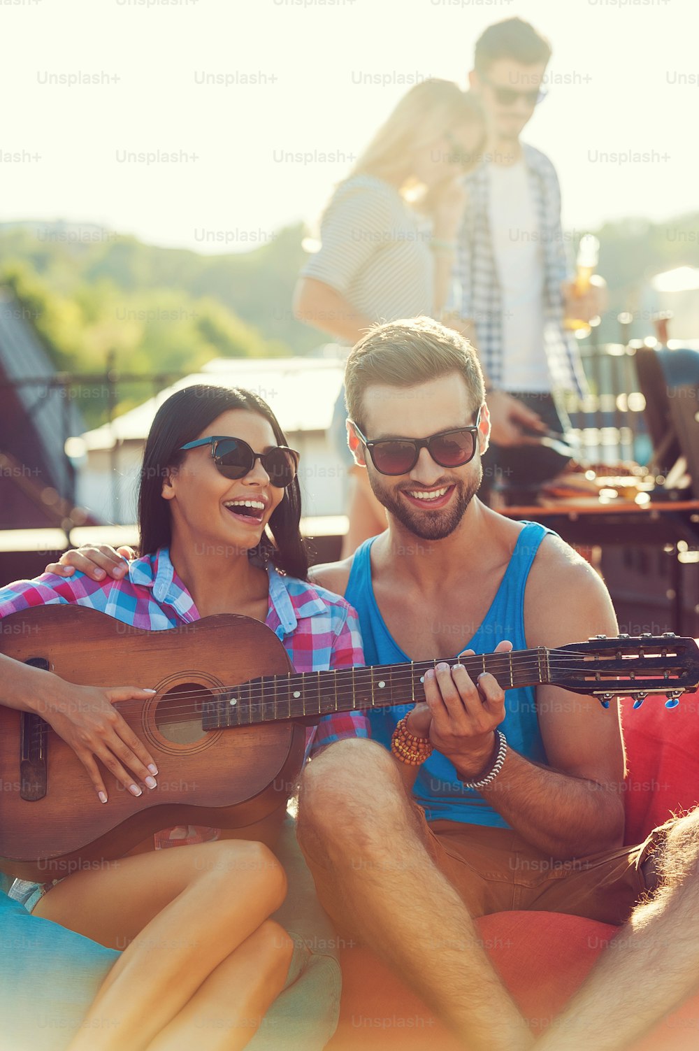 Joyful young man teaching his girlfriend to play the guitar while two people barbecuing in the background
