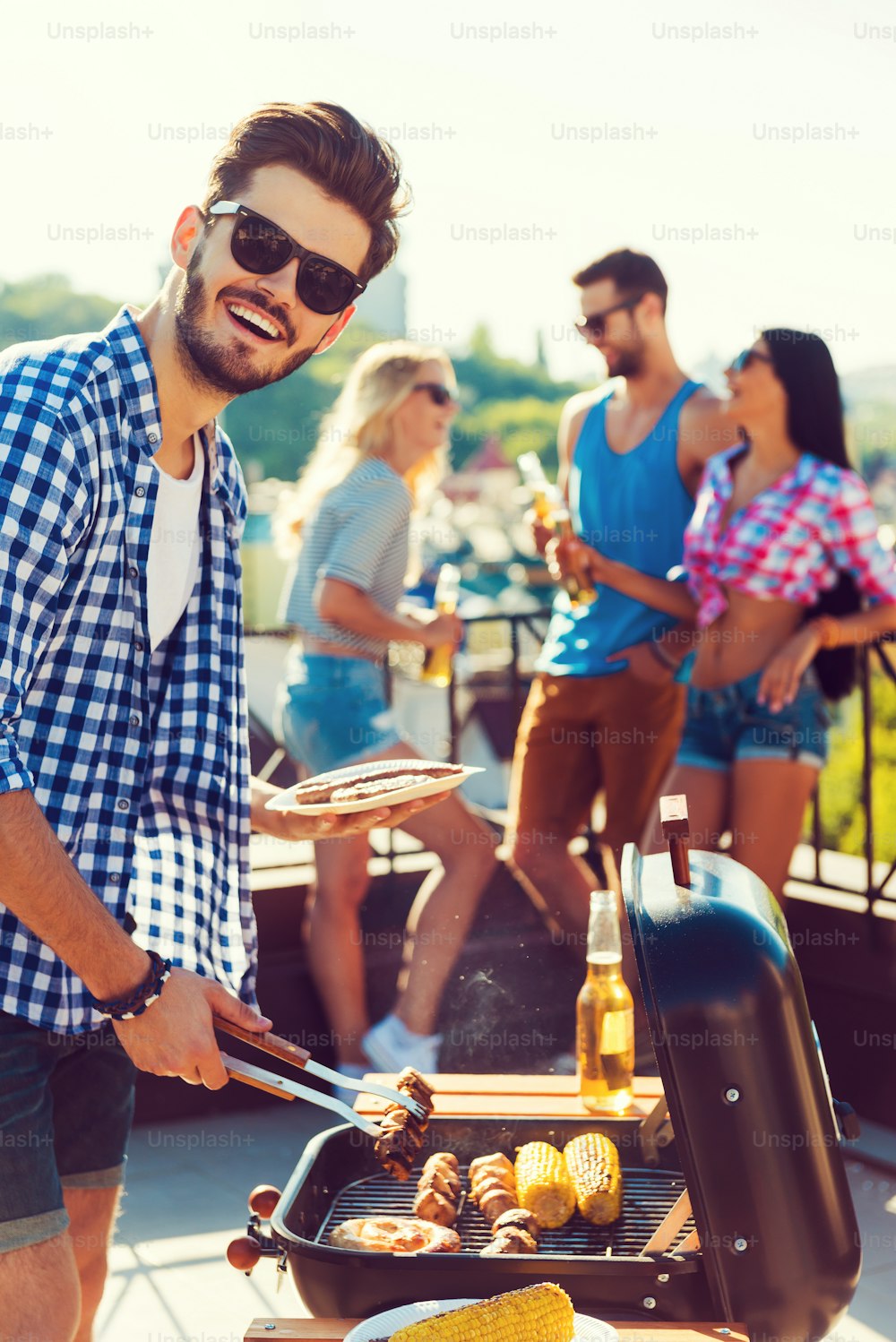 Happy young man barbecuing and looking at camera while three people having fun in the background