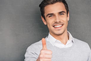Close-up of cheerful young man reaching out his hand in thumb up and smiling while standing against grey background
