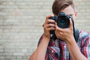 Confident young man focusing at you with his digital camera while standing against brick wall