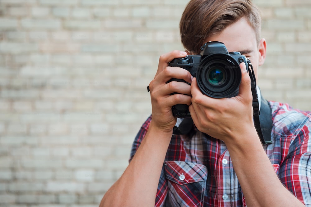 Confident young man focusing at you with his digital camera while standing against brick wall