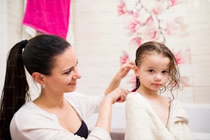 Beautiful young mother combing hair of her little daughter after taking bath